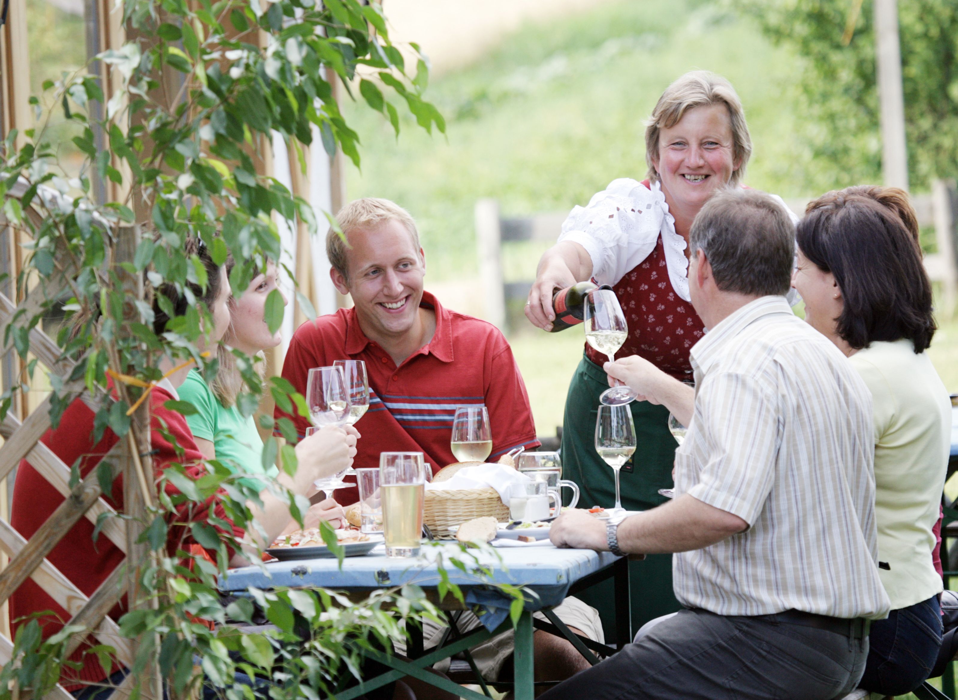 Eine Gruppe von Menschen sitzt an einem Tisch im Freien und genießt Wein. Eine Frau schenkt Wein ein, alle lächeln.