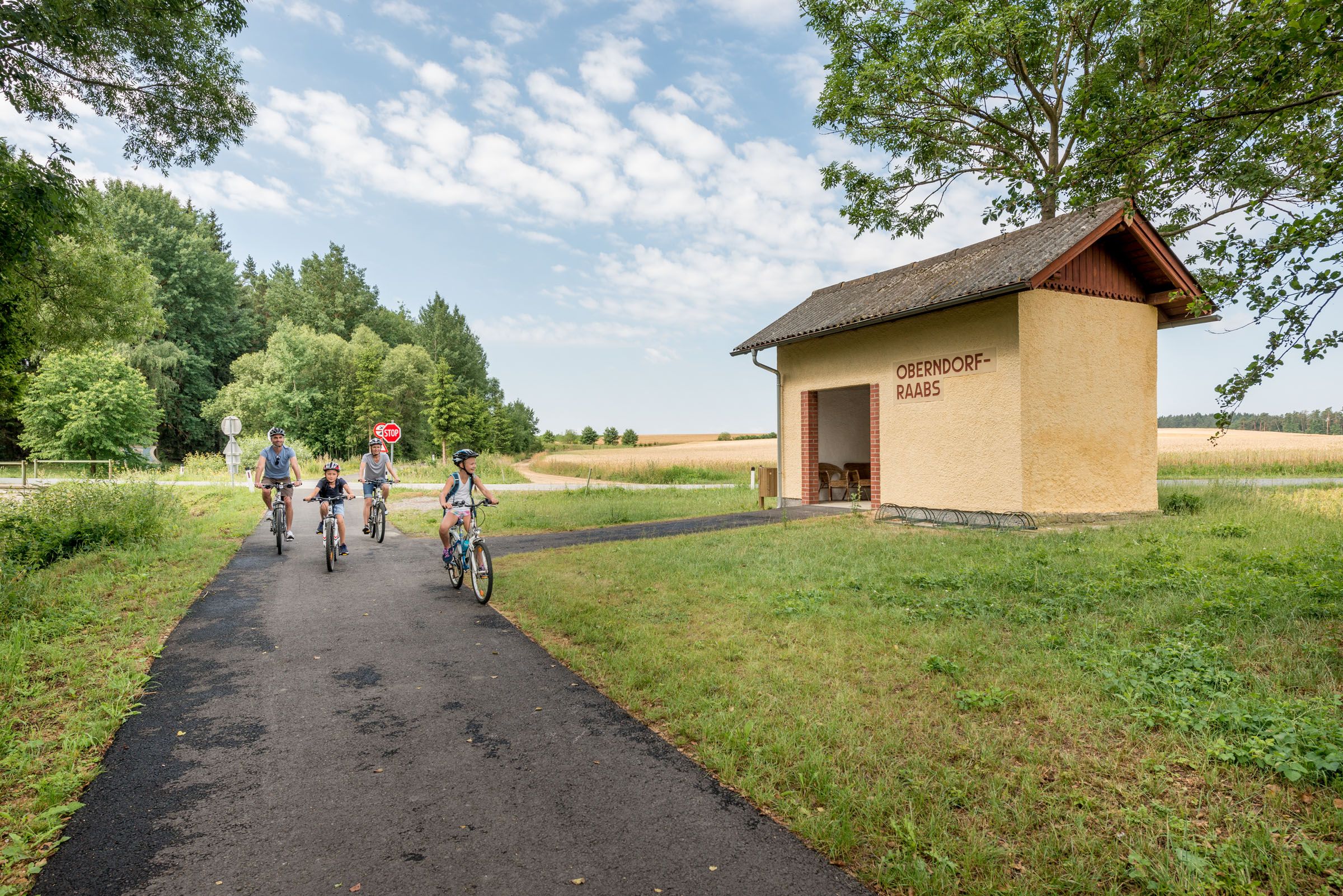 Eine Familie fährt mit Fahrrädern auf einem Weg neben einem gelben Gebäude mit der Aufschrift 'Oberndorf-Raabs'.