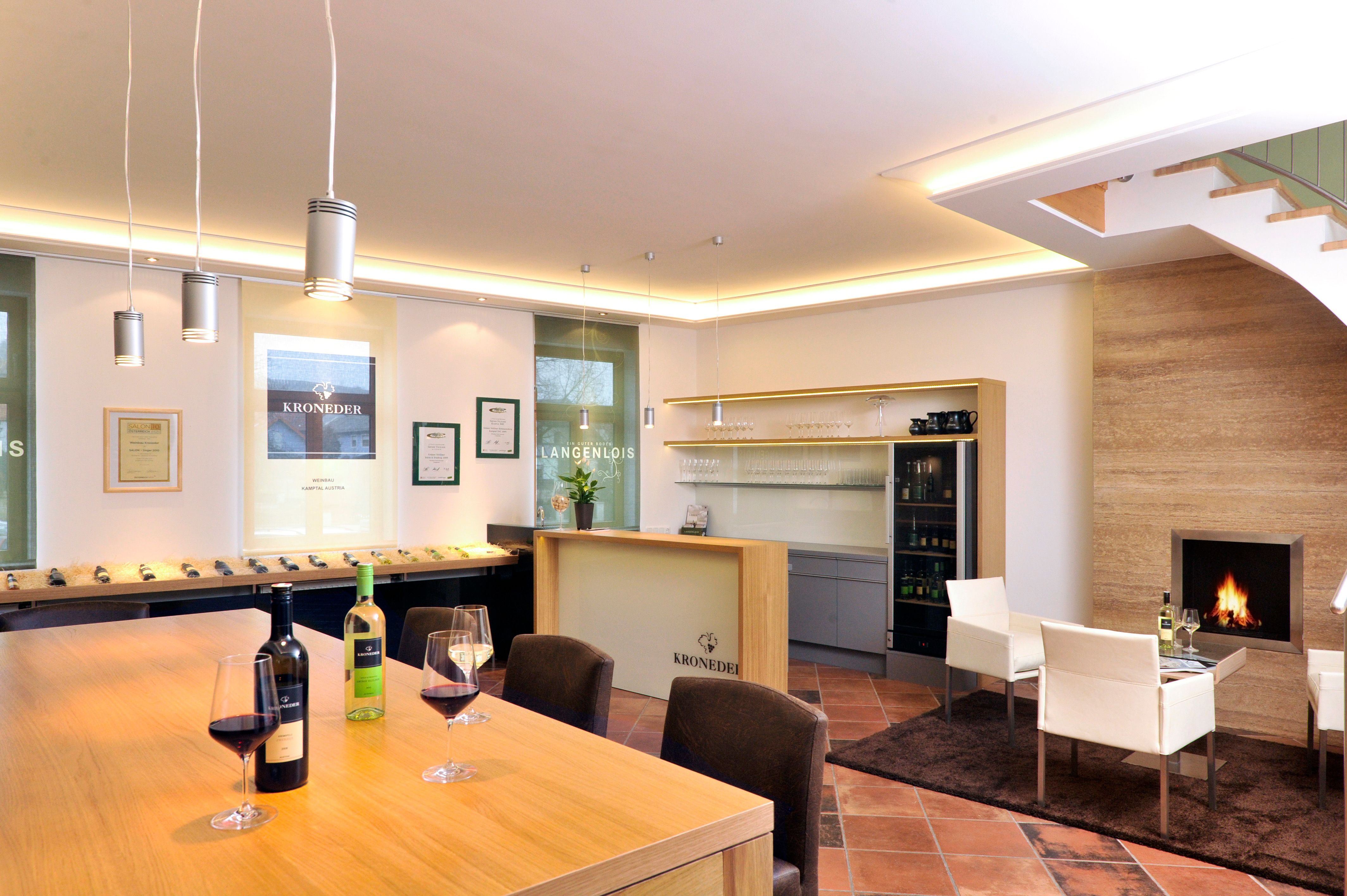 Innenansicht eines modernen Weinverkostungsraums mit Holztisch, Weingläsern, Kamin und Regalen.