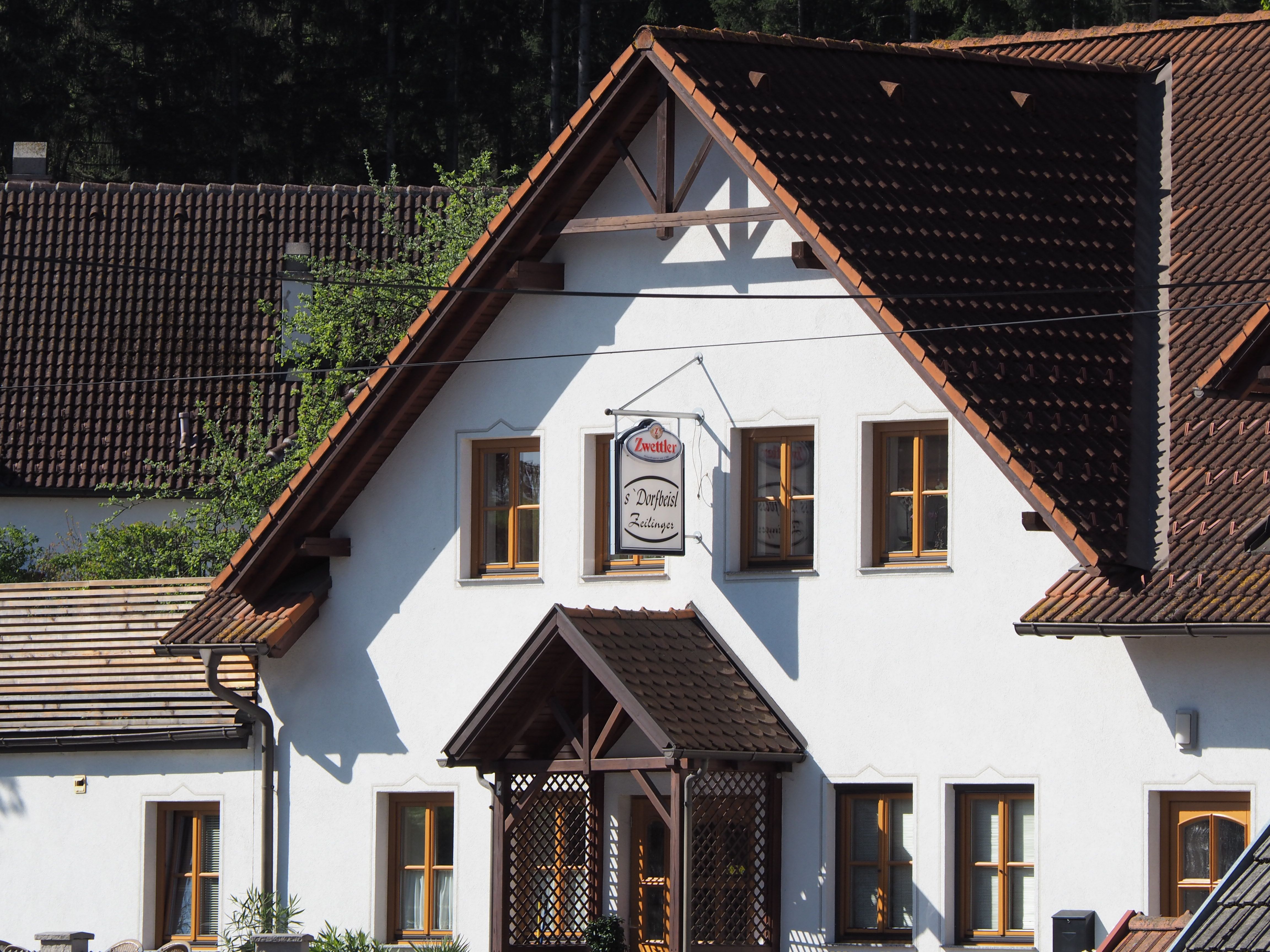 Ein traditionelles Gebäude mit weißen Wänden und braunen Dachziegeln. Ein Schild mit der Aufschrift 'Zwettler Dorfwirt Zeilinger' hängt an der Fassade.