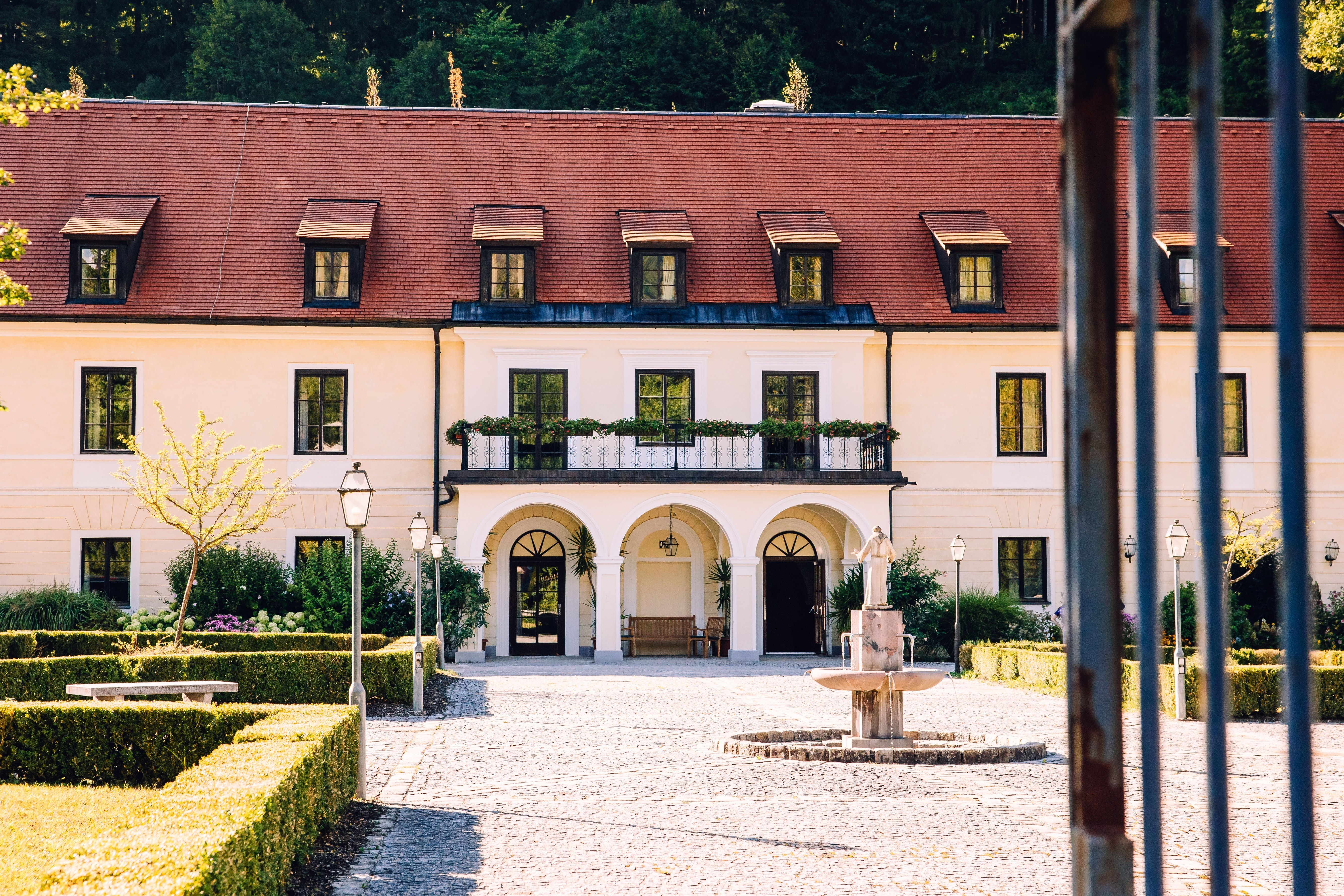 Innenhof der Kartause Gaming mit rotem Dach, Brunnen und gepflegten Hecken; im Vordergrund ein Tor mit Stäben.