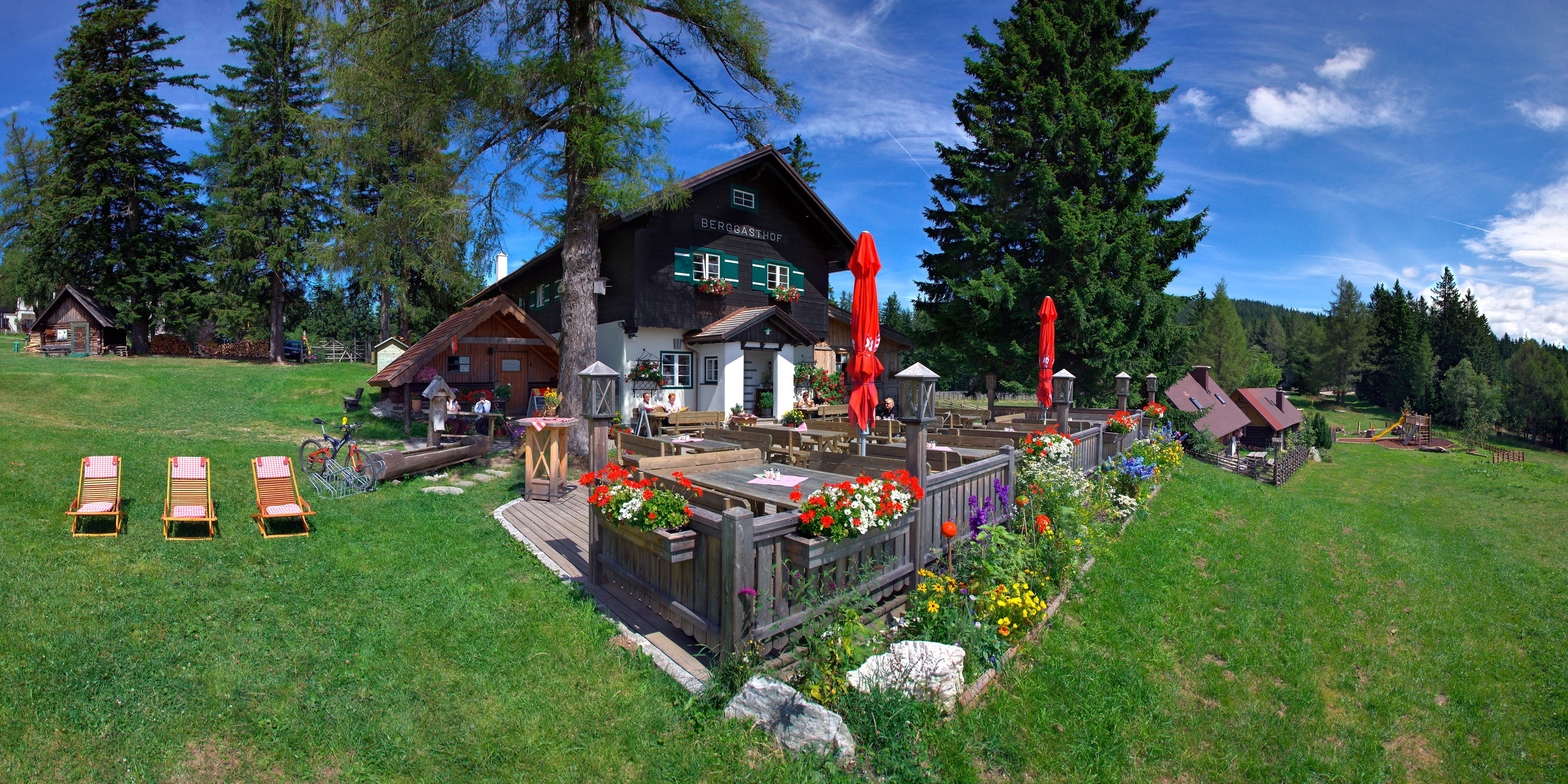 Ein Berggasthof mit Terrasse, Blumen und Liegestühlen auf einer grünen Wiese, umgeben von Bäumen und einem klaren blauen Himmel.