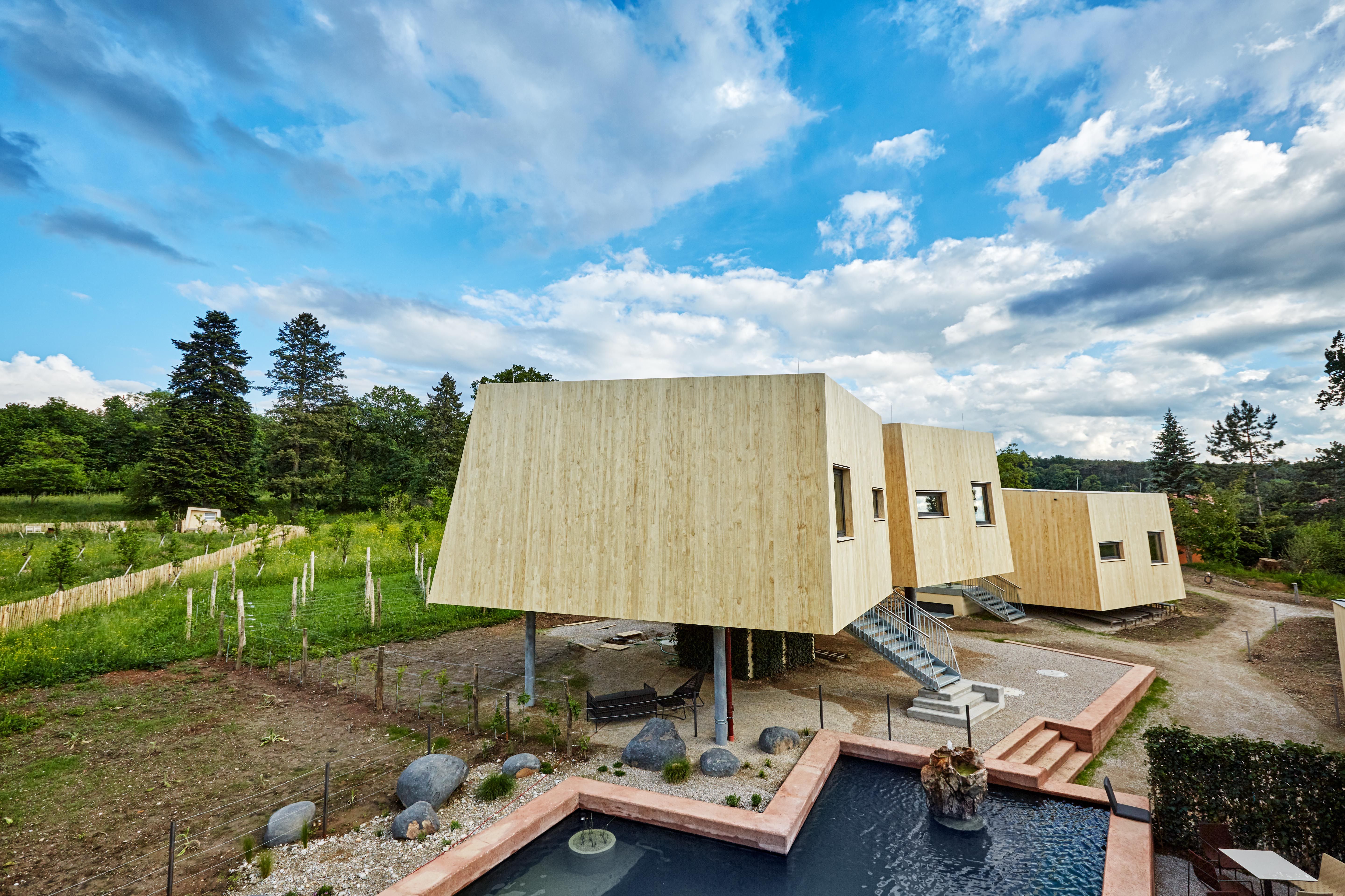 Moderne Gartenlofts aus Holz mit Treppen, umgeben von Natur und einem Teich im Vordergrund.