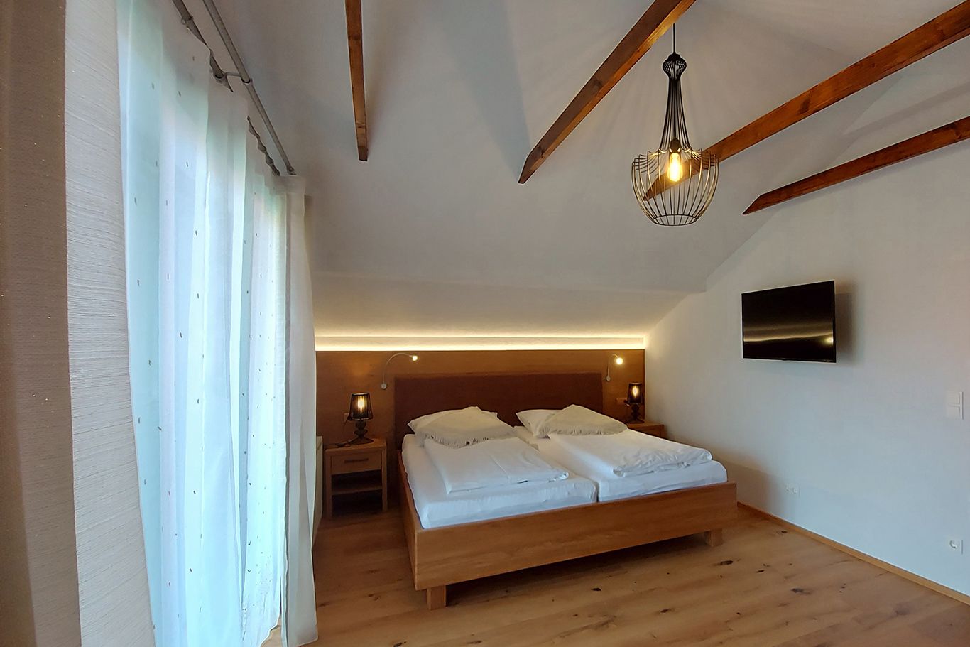 Doppelzimmer mit Holzbalken, Doppelbett, Nachttischen, Lampe und Fernseher an der Wand.