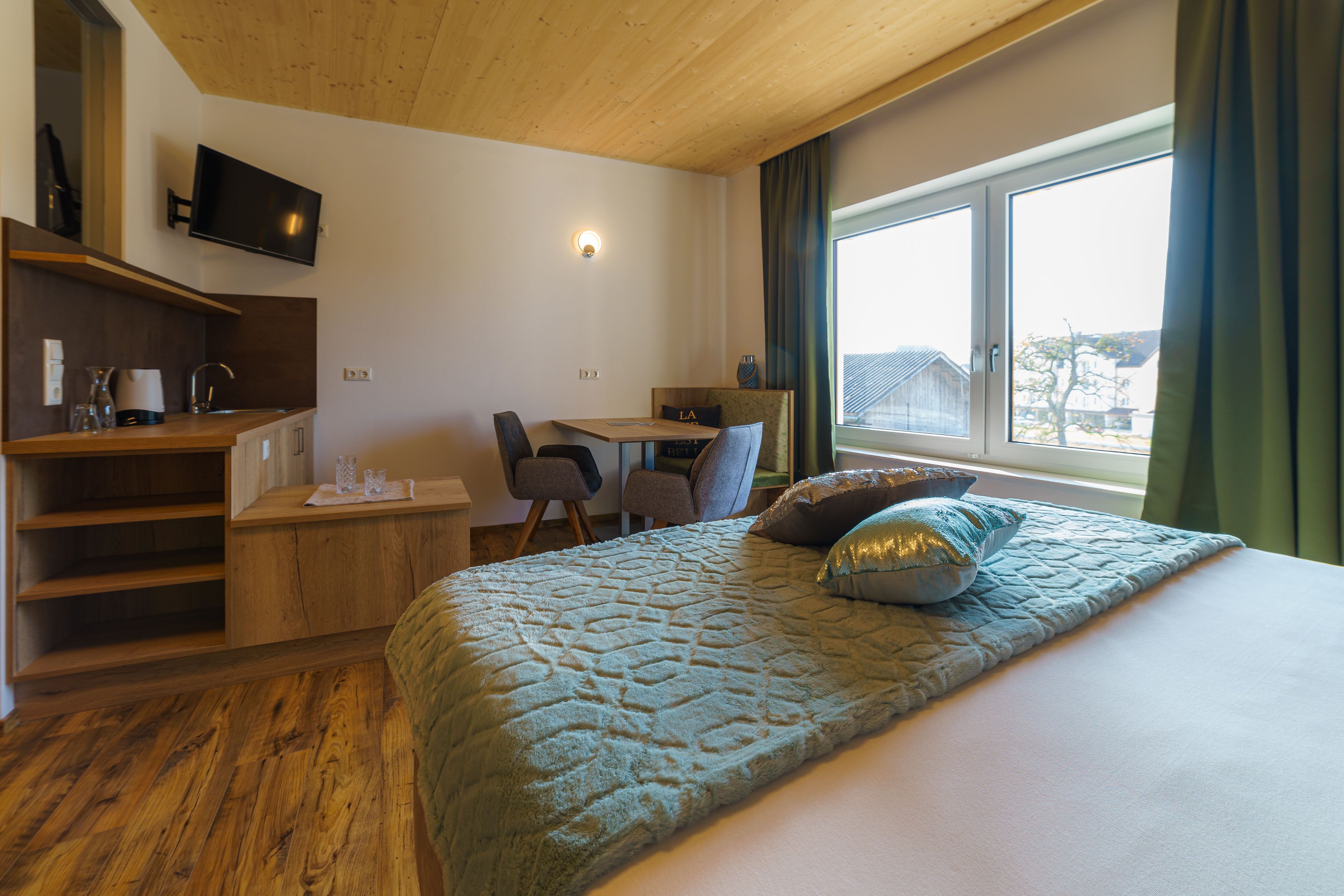Gemütliches Hotelzimmer mit Holzboden, Doppelbett, kleinem Tisch und Stühlen, Fernseher und großem Fenster mit Ausblick.