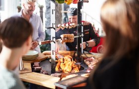 Ein Grillmeister bereitet Fleisch und Gem&uuml;se auf einem offenen Grill vor, umgeben von interessierten Zuschauern.