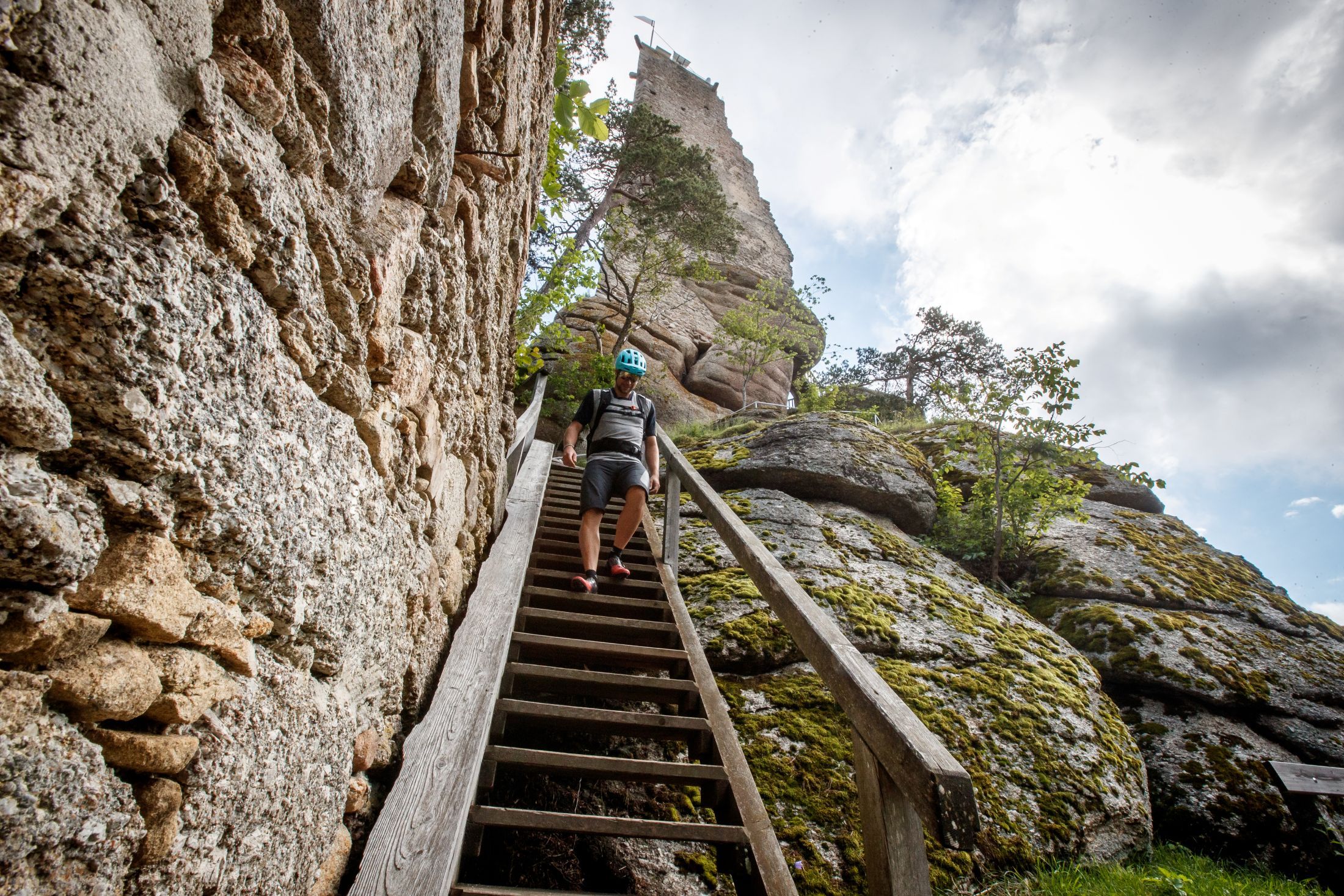 Ein Mann mit Helm geht eine Holztreppe hinunter, neben einer steinernen Burgruine und Felsen.