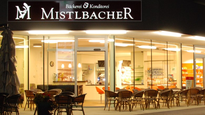 Außenansicht eines Cafés mit dem Namen Mistelbacher, beleuchtet und mit Sitzgelegenheiten im Freien.