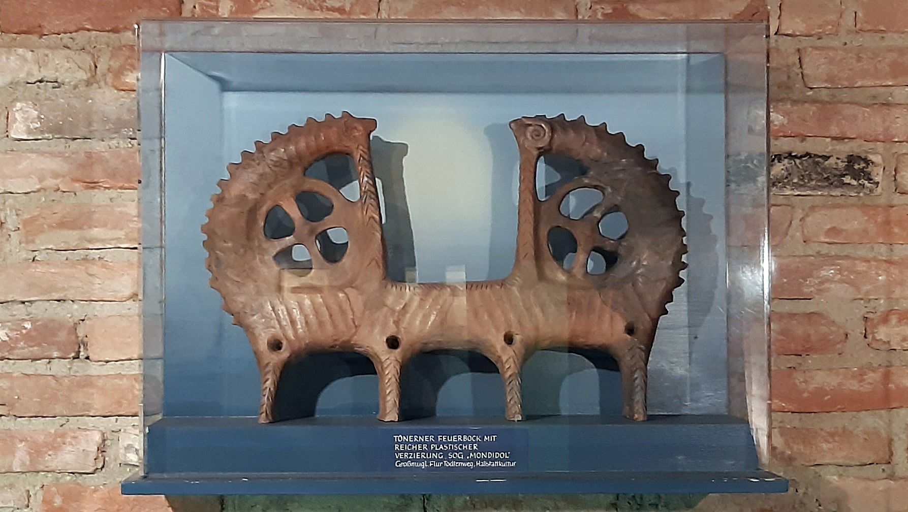 Tonfigur eines Feuerbocks in einer Vitrine vor einer Ziegelwand.