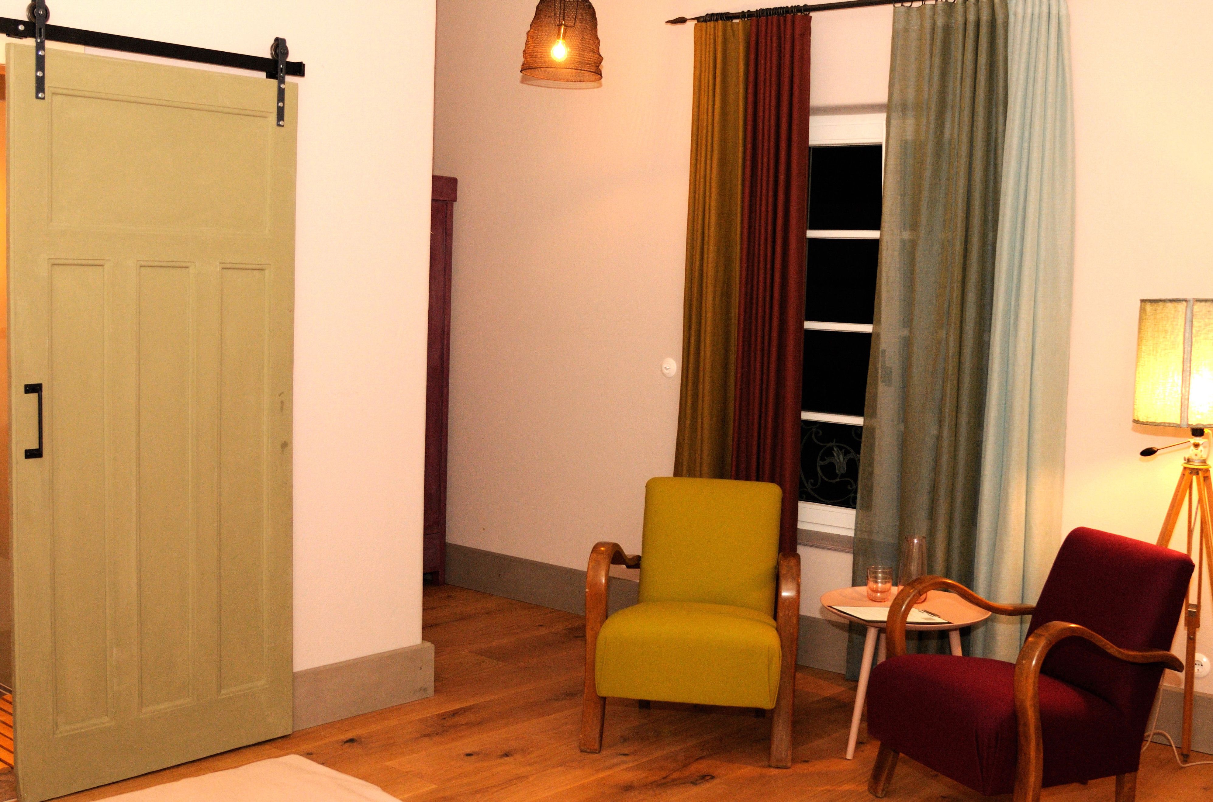 Ein gemütliches Zimmer mit zwei Sesseln, einem kleinen Tisch und einer Lampe. Ein Schiebetür und bunte Vorhänge sind ebenfalls zu sehen.