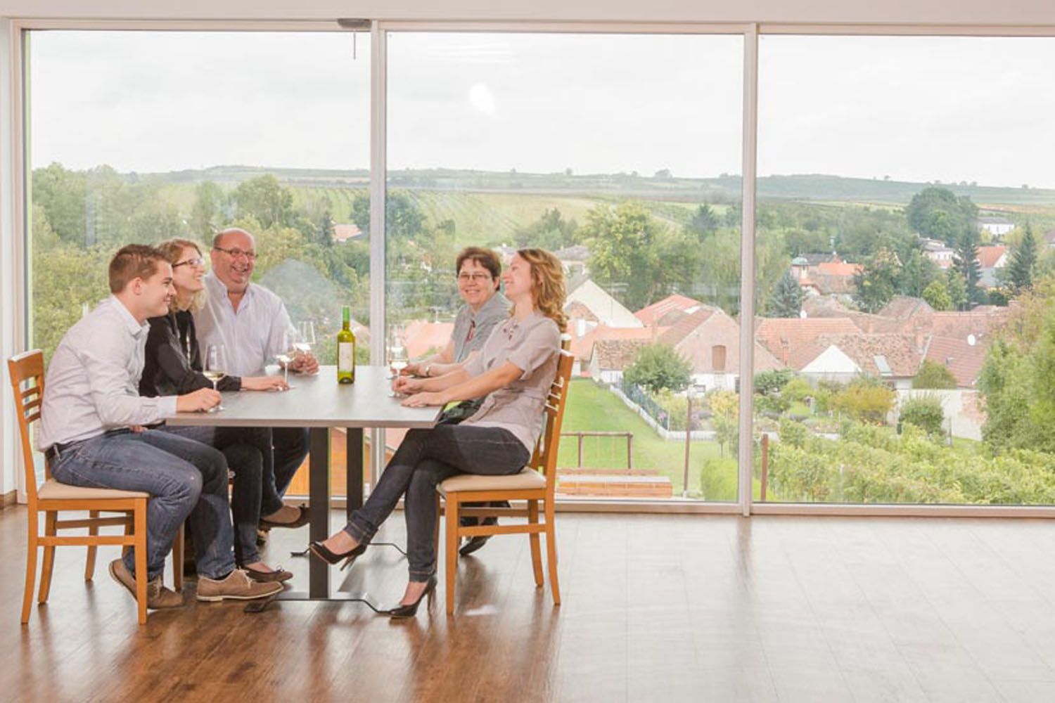 Eine Familie sitzt an einem Tisch mit Wein, im Hintergrund eine Landschaft mit Häusern und Feldern.
