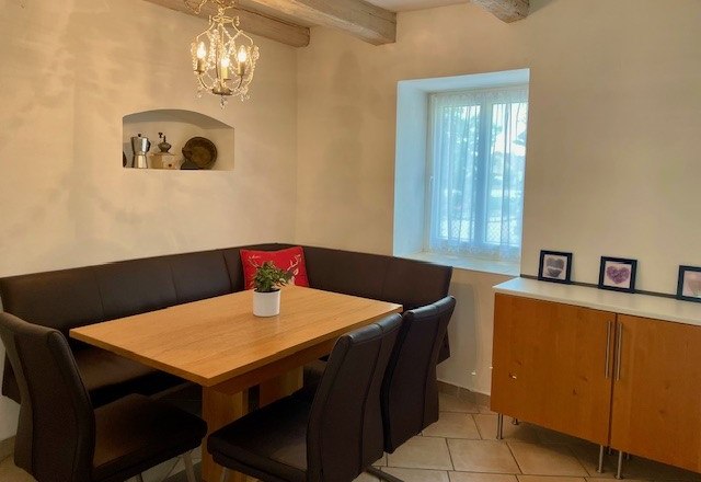 Ein gem&uuml;tlicher Essbereich mit Holztisch, schwarzen St&uuml;hlen und Eckbank. An der Wand h&auml;ngt ein Kronleuchter, daneben ein Fenster und ein Sideboard.