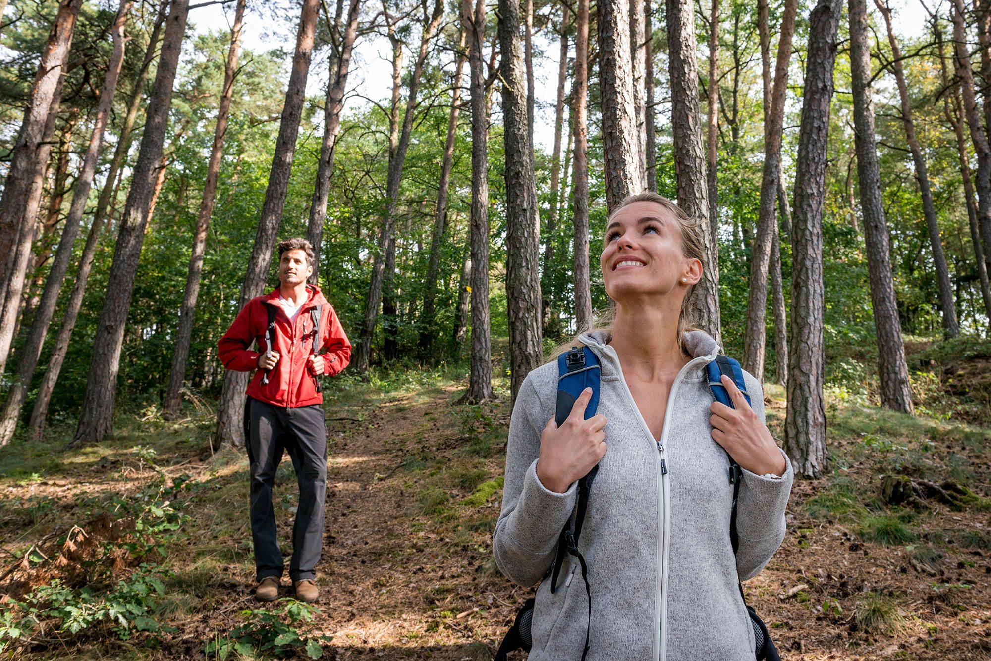 Zwei Personen wandern im Wald, eine Frau im Vordergrund lächelt, ein Mann im Hintergrund trägt eine rote Jacke.