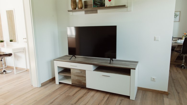 Wohnzimmer mit Fernseher auf einem weißen TV-Schrank, darüber ein Regal mit Dekorationen.