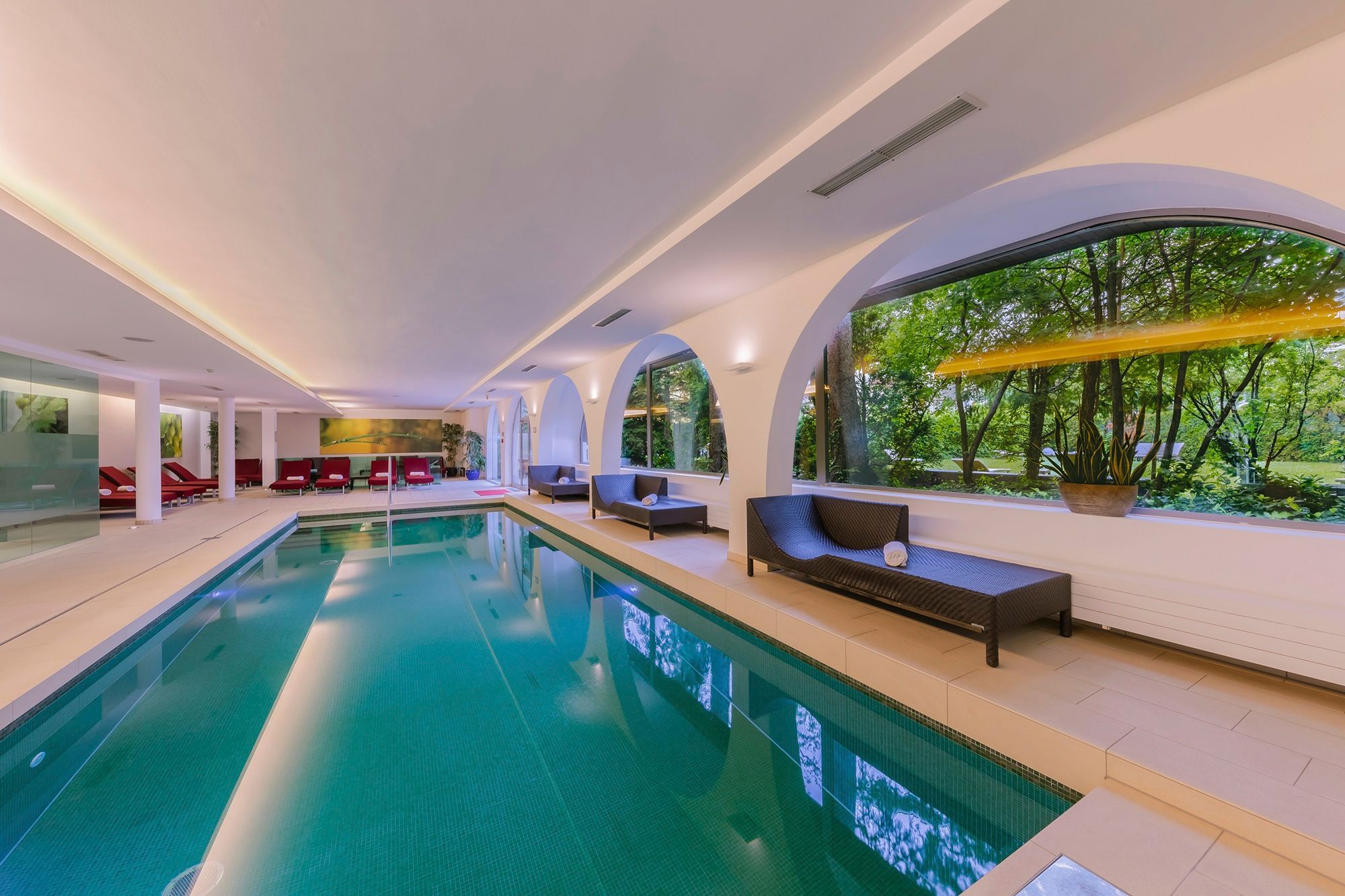 Innenansicht eines eleganten Spa-Bereichs mit Pool, Liegen und großen Rundbogenfenstern zum Garten.