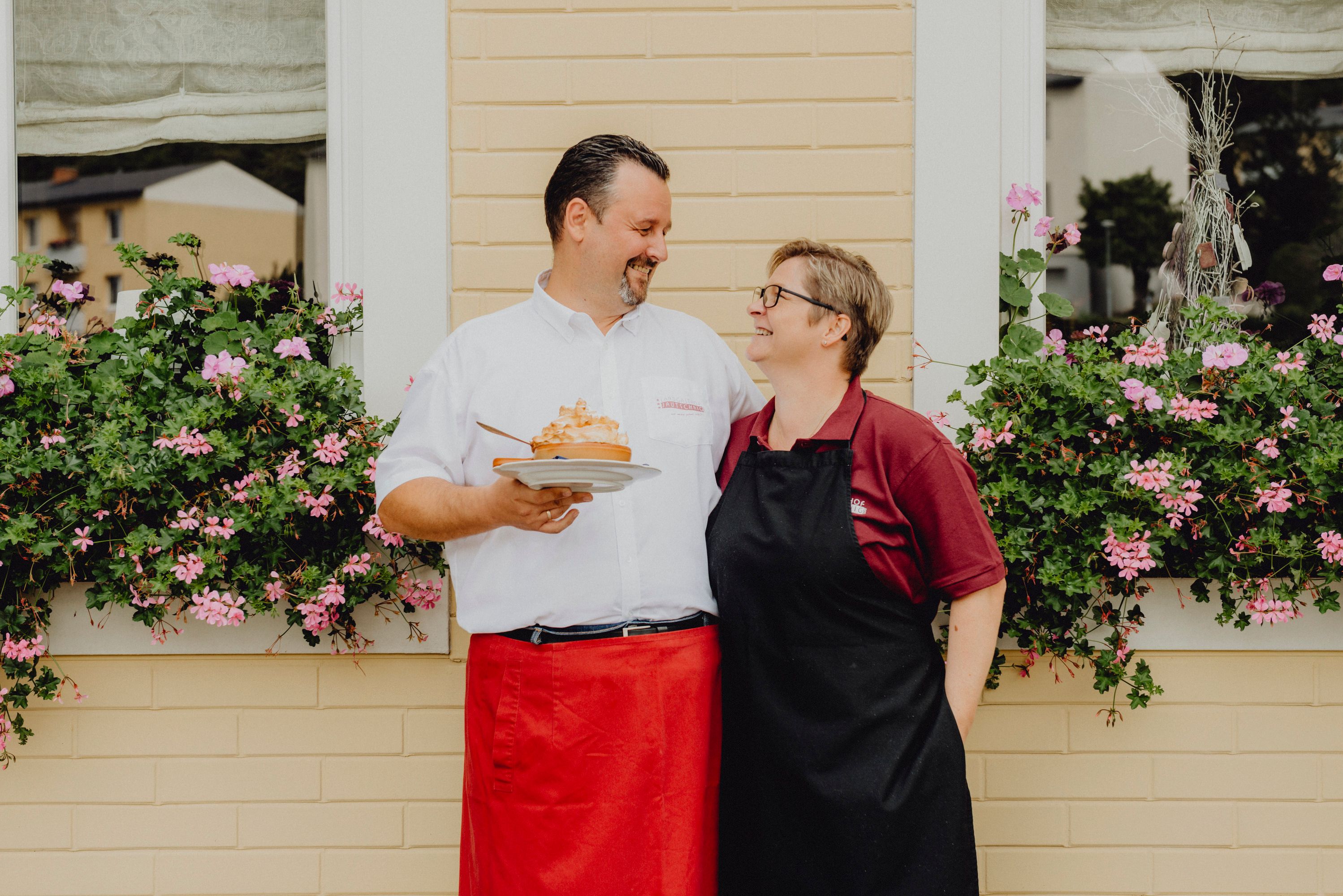Ein Mann und eine Frau stehen lächelnd vor einem Fenster mit Blumen.