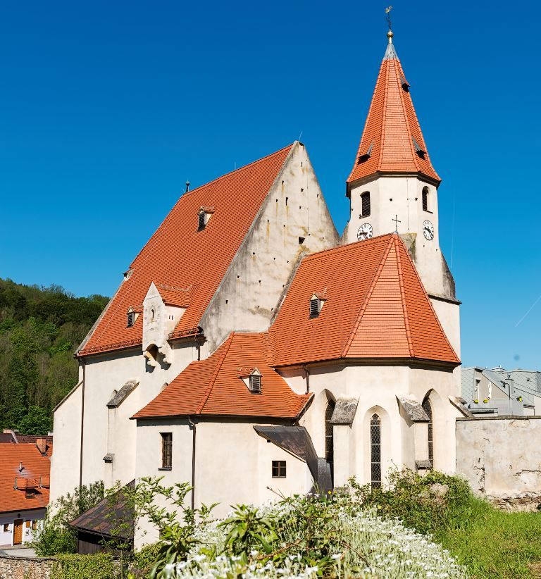 Wehrkirche Edlitz mit vielen Seitenbereichen, roten Ziegeldächern, einem spitzen Turm und blauem Himmel.