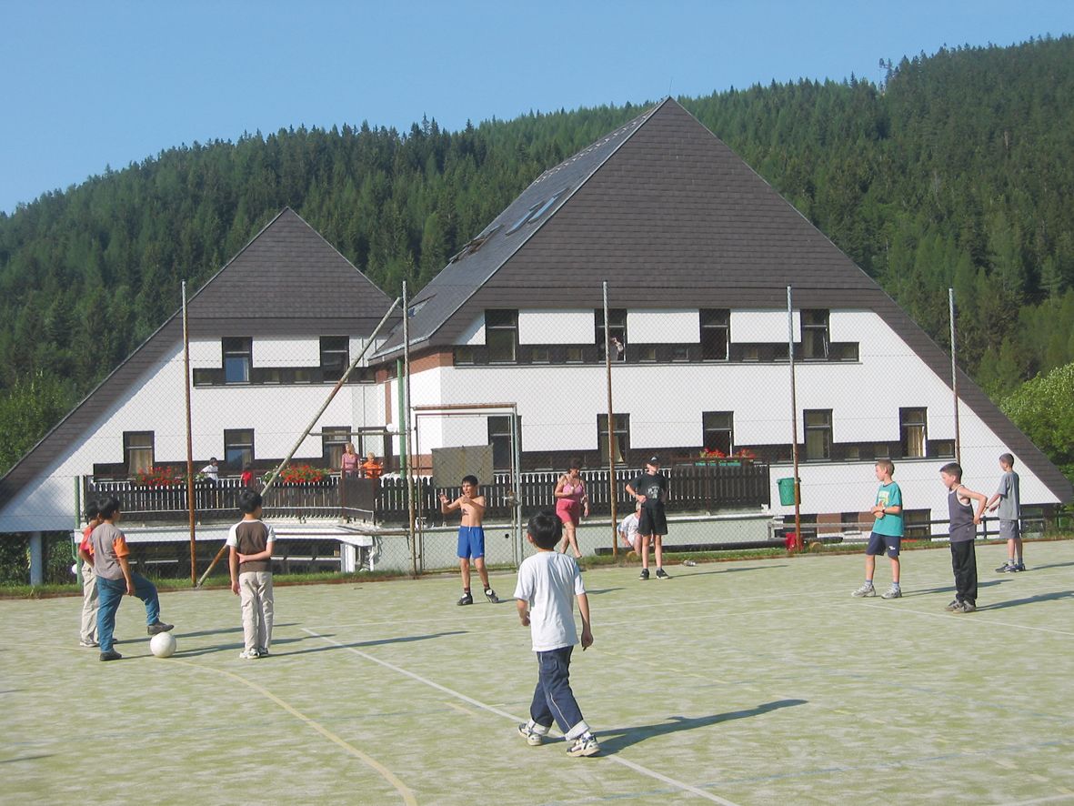 Kinder spielen Fußball auf einem Sportplatz vor einem Hotel in einer bewaldeten Gegend.
