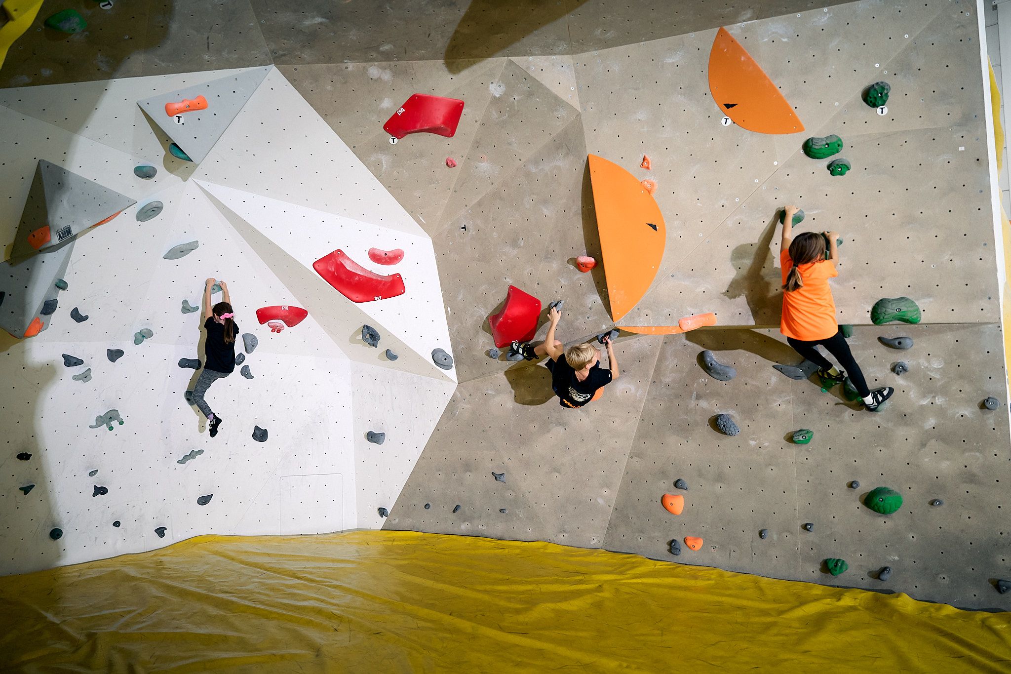 Drei Kinder klettern an einer Indoor-Boulderwand mit bunten Griffen.