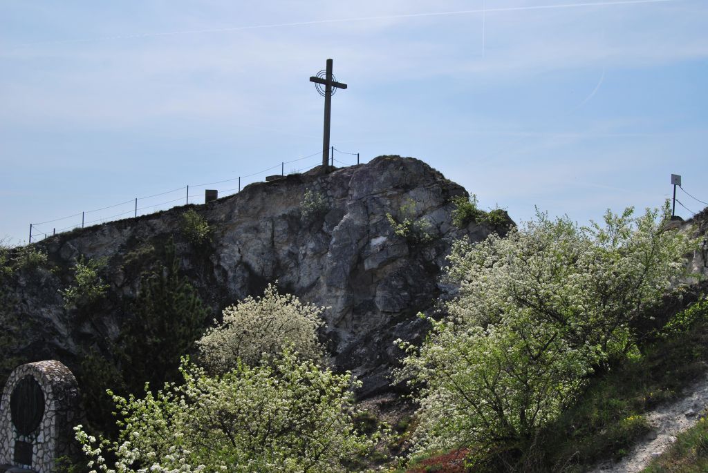 Ein Kreuz steht auf einem Hügel mit blühenden Bäumen im Vordergrund.
