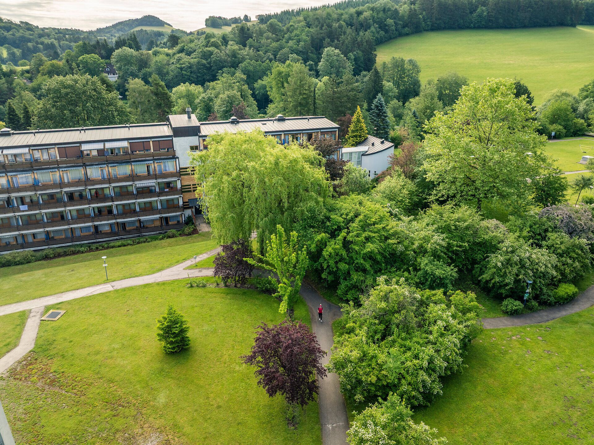 Luftansicht auf das Gesundheitsresort Königsberg und Garten mit grüner Wiese, Sträuchern und Bäumen. Zwischen den Bäumen ist auf einem Weg eine Person erkennbar.