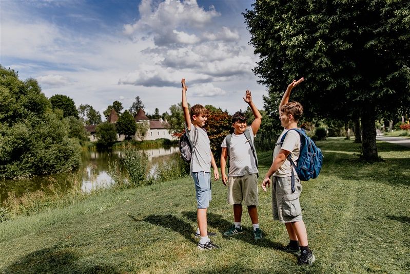 Drei Jungen mit Rucksäcken stehen auf einer Wiese neben einem Teich und heben die Arme in die Luft.