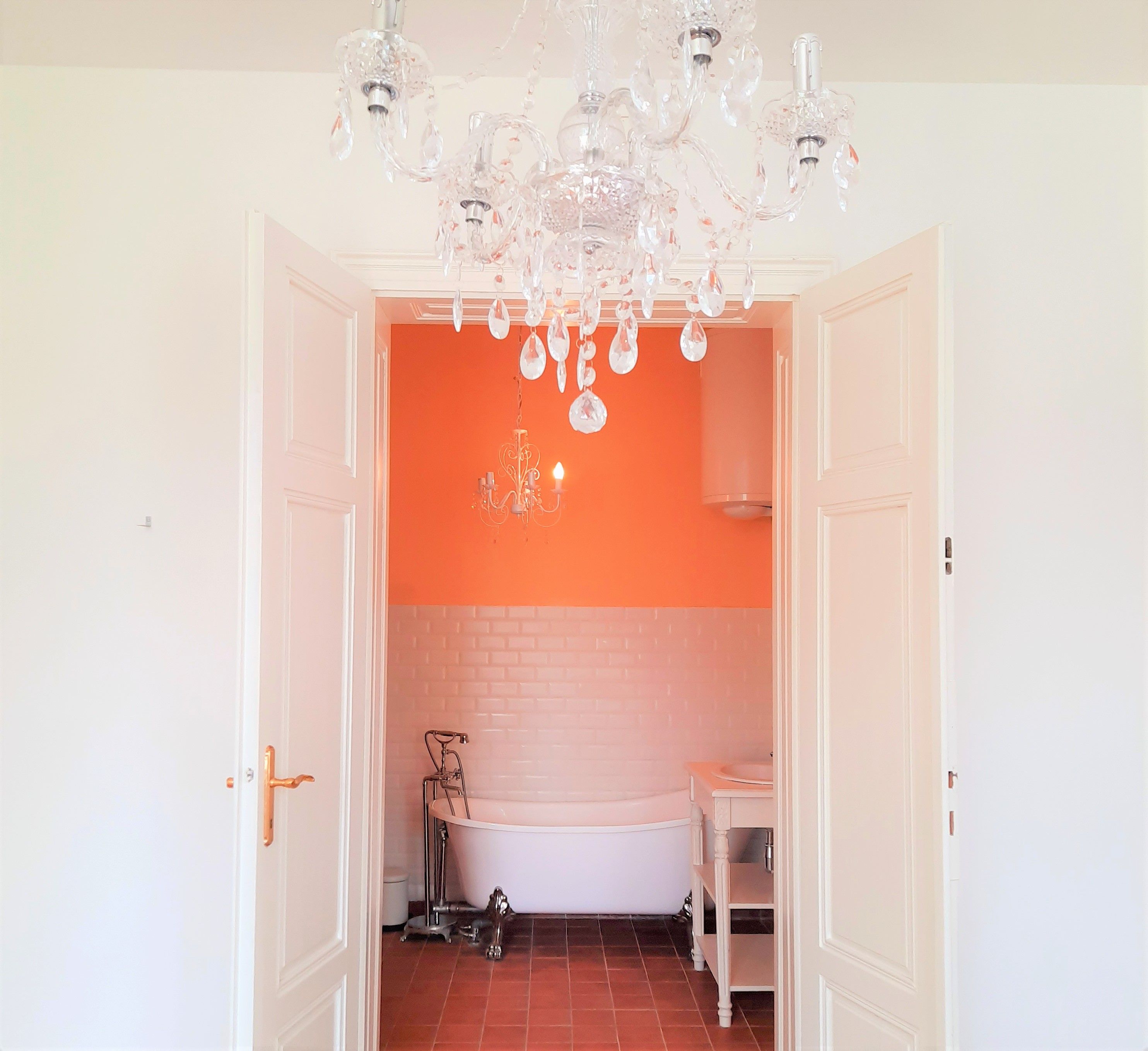 Blick in ein Badezimmer mit freistehender Badewanne, orangefarbener Wand und Kronleuchter.