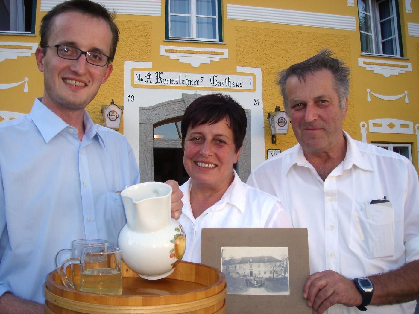 Drei Personen vor einem gelben Gasthaus mit einem Tablett und einem alten Foto.