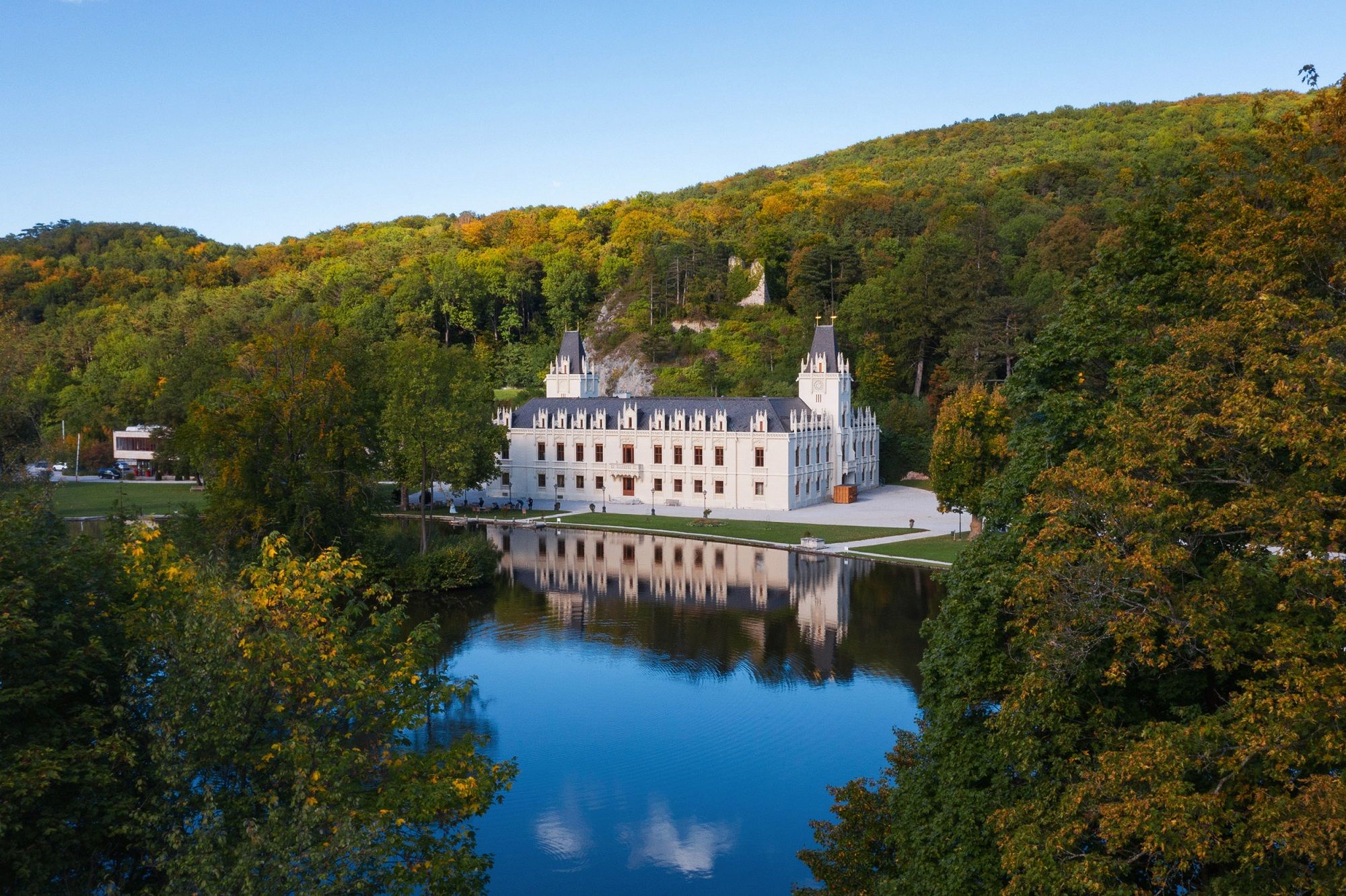 Schloss Hernstein mit Spiegelung im Wasser, umgeben von Bäumen und Hügeln.