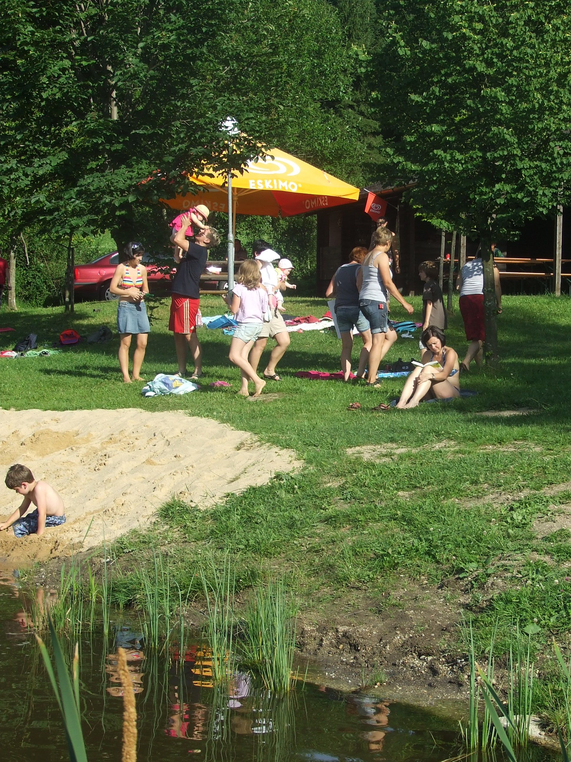 Menschen entspannen an einem Naturbadeteich mit Sonnenschirm und Bäumen.