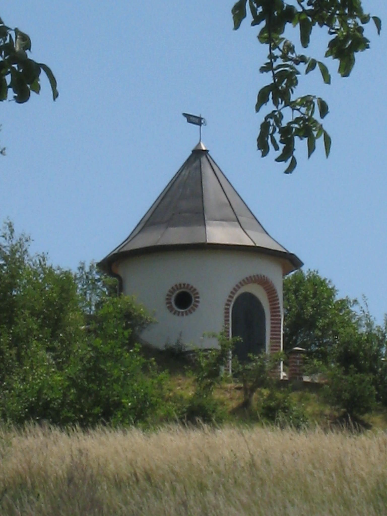 Ein runder Wasserturm mit spitzem Dach und Wetterfahne, umgeben von Bäumen und Wiese.