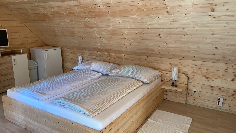 Ein gem&uuml;tliches Doppelzimmer mit Holzverkleidung, einem Doppelbett, einem kleinen K&uuml;hlschrank und einem Fernseher auf einer Kommode.