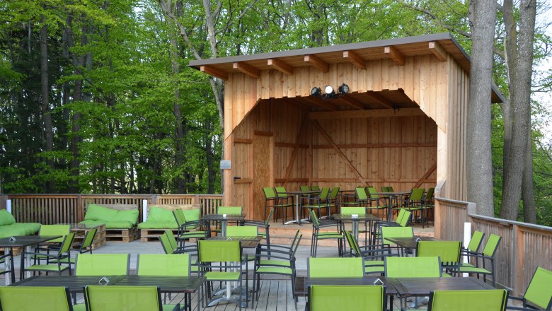 Eine Holzterasse mit schwarzen Tischen und gr&uuml;nen St&uuml;hlen und einer &uuml;berdachten Holzb&uuml;hne.