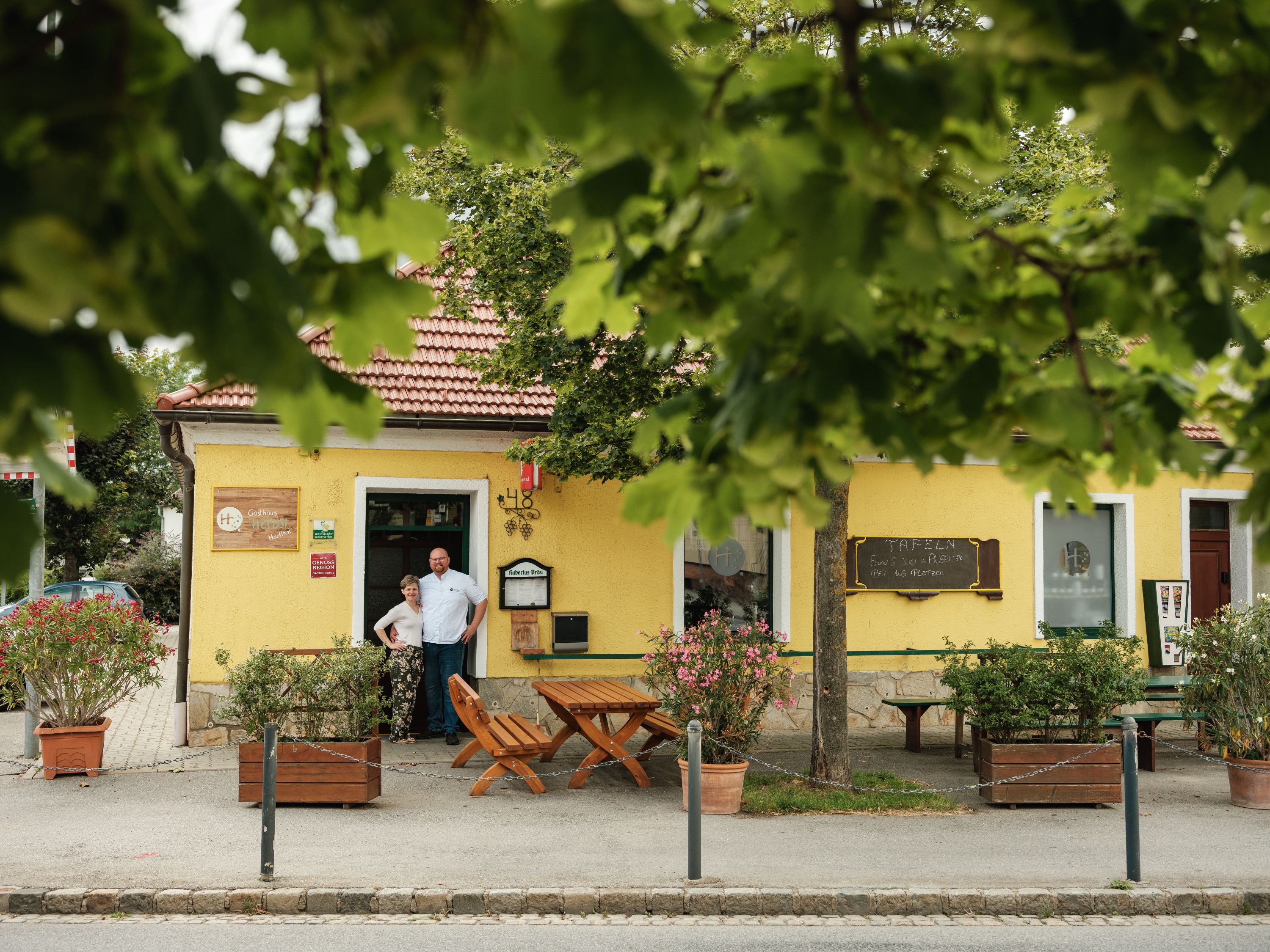 Ein gelbes Gasthaus mit rotem Dach, umgeben von Bäumen. Zwei Personen stehen vor dem Eingang.