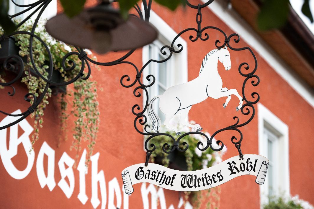 Metallschild mit weißem Pferd und Schriftzug 'Gasthof Weißes Rößl' vor rotem Gebäude.