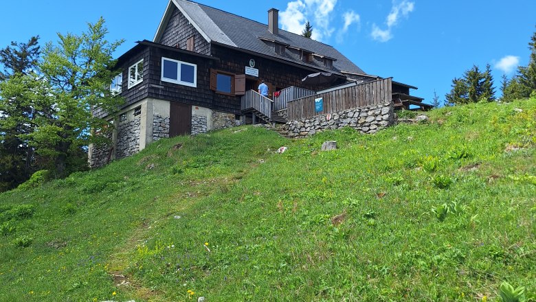 Waldfreundehütte Obersberg, © Stefanie Gaulhofer Eine Berghütte auf einer grünen Wiese unter blauem Himmel.