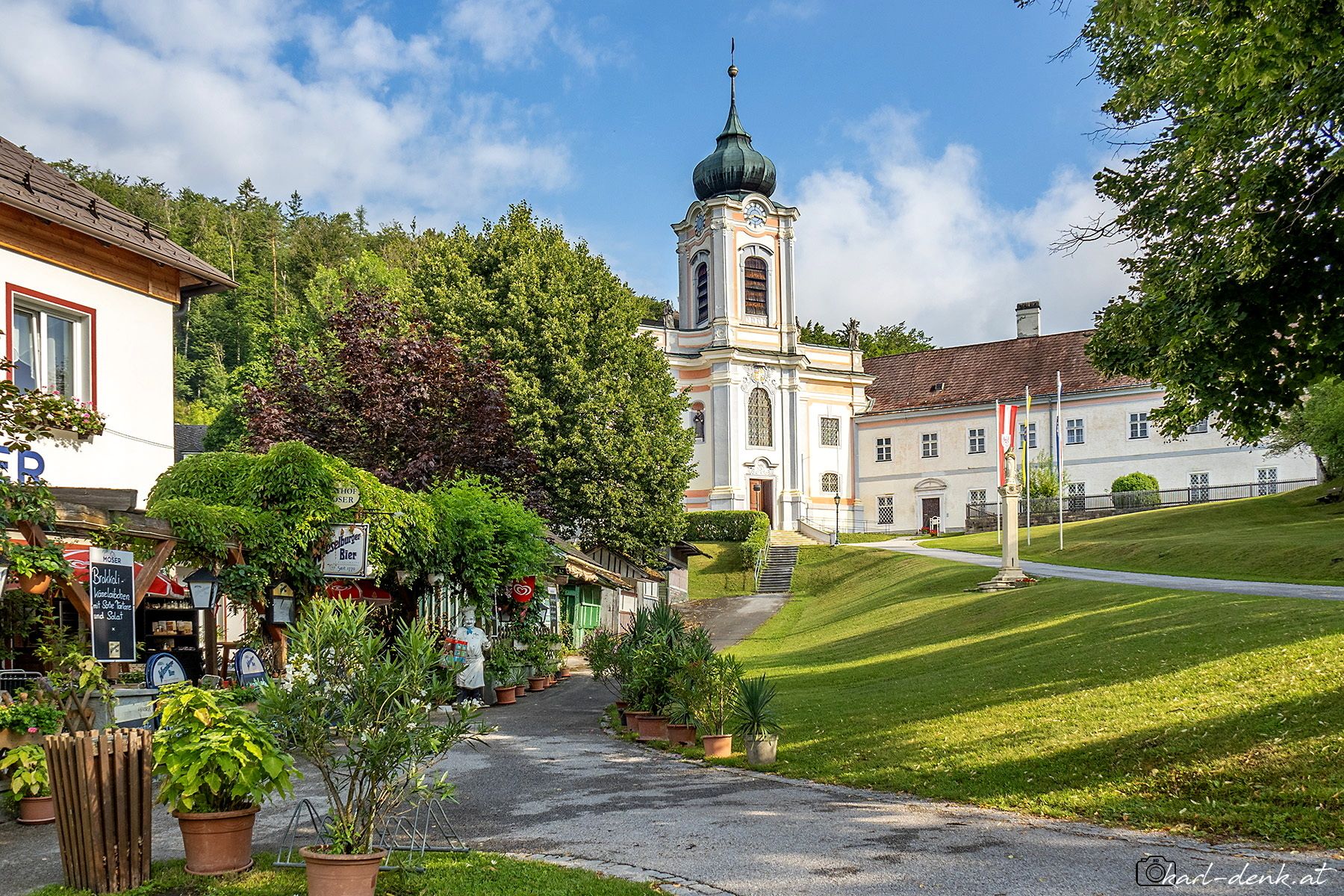 Ein malerisches Dorf mit einer Kirche im Barockstil und einem Gasthaus im Vordergrund, umgeben von grünen Bäumen und einem blauen Himmel.