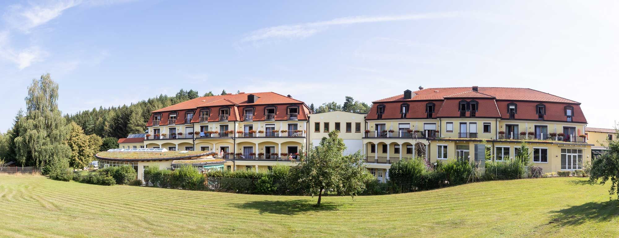 Außenansicht des Gesundheitshotels Moorbad Bad Großpertholz mit rotem Dach und Balkonen, umgeben von grüner Landschaft.