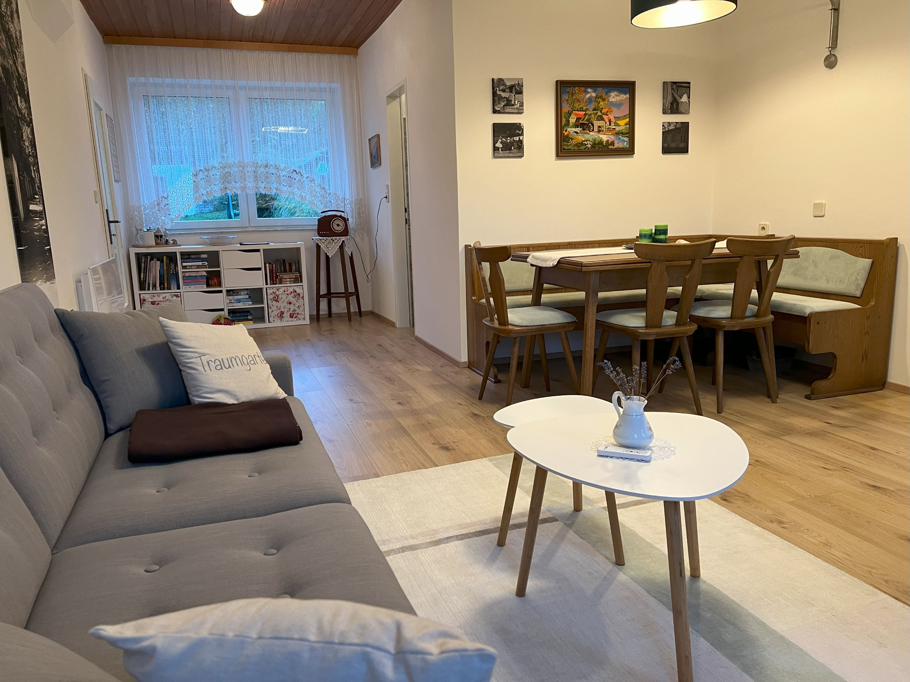 Gemütliches Wohnzimmer mit grauem Sofa, Esstisch und Stühlen, Bücherregal und Fenster mit Gardinen.