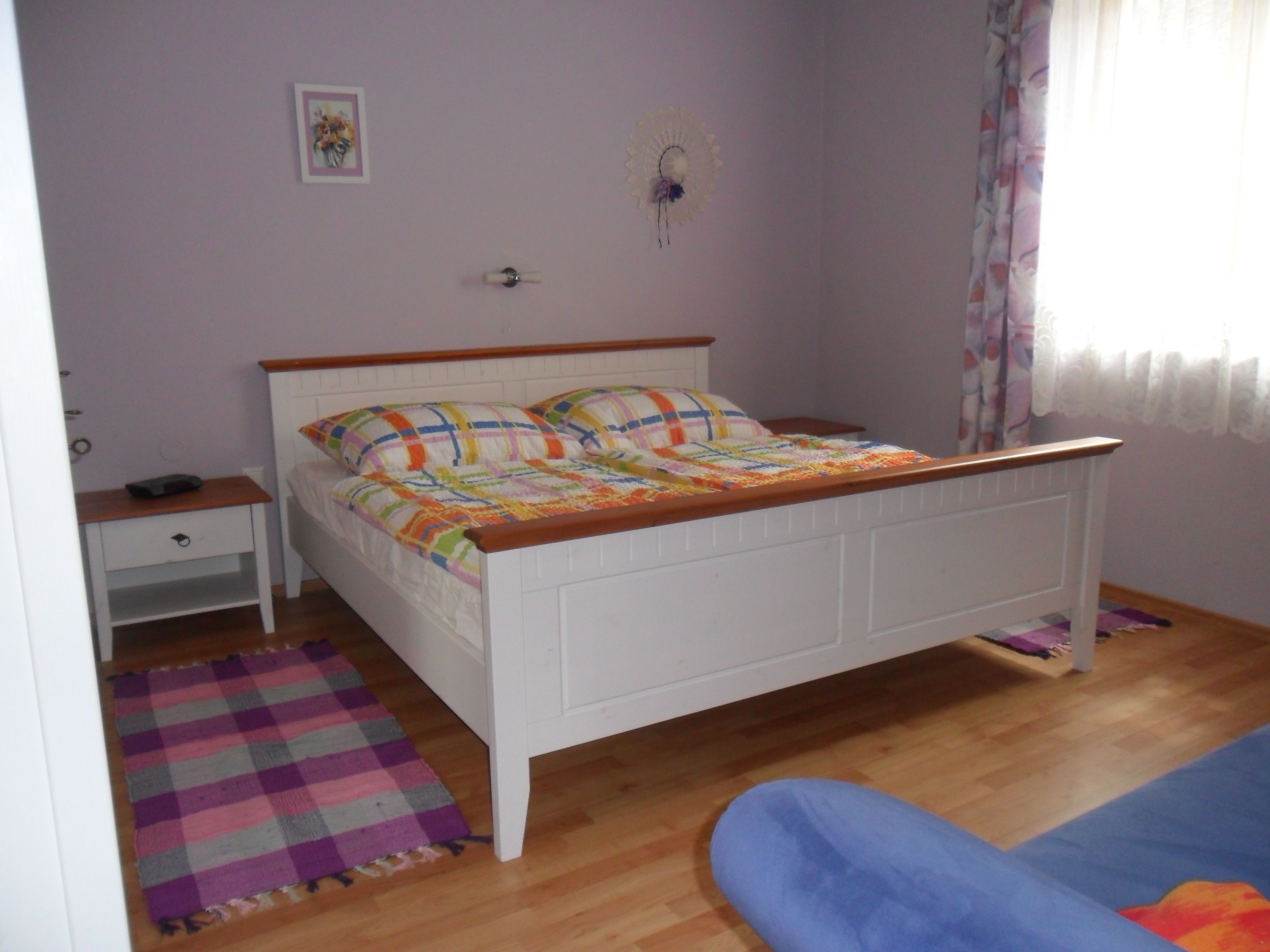 Ein Schlafzimmer mit einem weißen Bett, bunten Bettdecken, einem Nachttisch und einem Fenster mit Vorhängen.