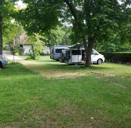 Zwei Wohnmobile stehen auf einem gr&uuml;nen Campingplatz unter B&auml;umen.