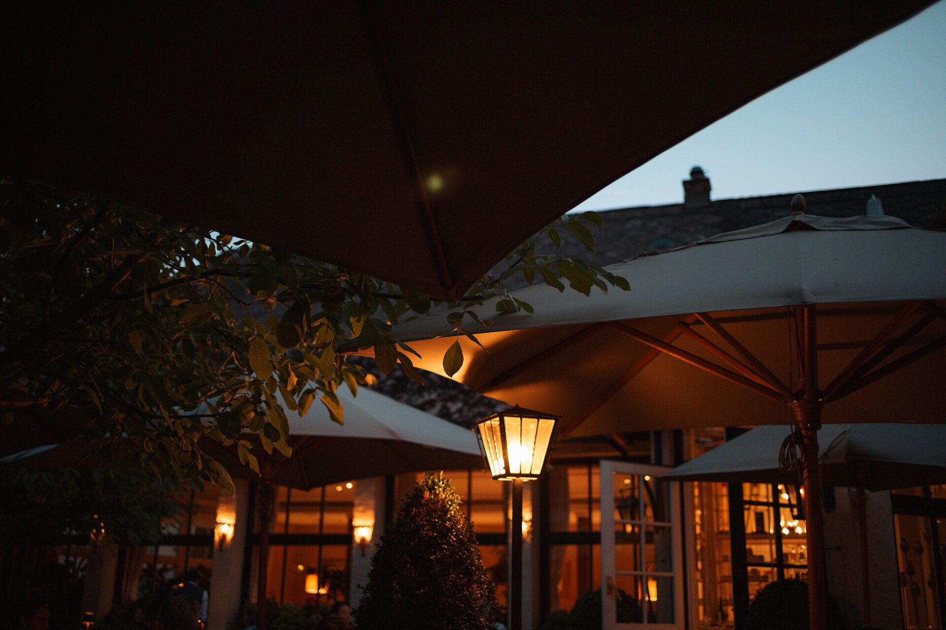 Die sanfte Abenddämmerung hüllt die Terrasse in ein warmes Licht, während die eleganten Sonnenschirme sanft im Wind wiegen. Gemütliche Tische laden dazu ein, die köstlichen Spezialitäten der Region zu genießen und den Tag in entspannter Atmosphäre ausklingen zu lassen.