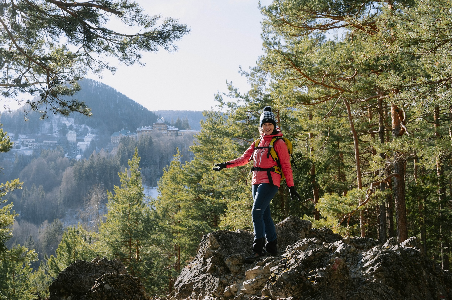 Person in pinker Jacke wandert auf einem felsigen Pfad im Wald mit Bergen im Hintergrund.