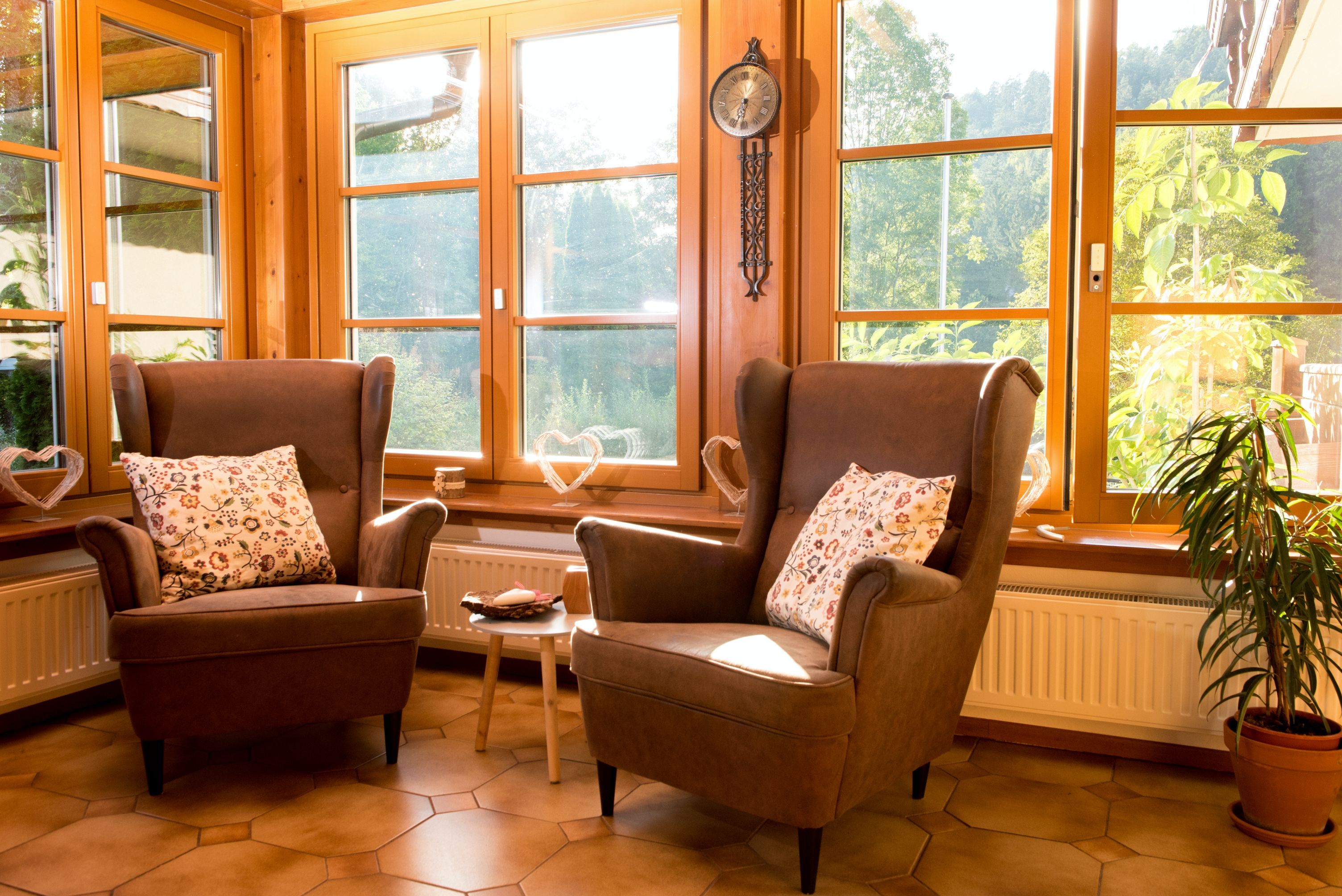 Zwei braune Sessel mit gemusterten Kissen vor großen Fenstern in einem sonnigen Raum.