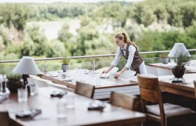 Eine Kellnerin deckt Tische auf einer Terrasse mit Blick auf den Nationalpark Donau-Auen.