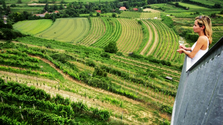 Frau mit Weinglas blickt von einem Aussichtspunkt auf gr&uuml;ne Weinberge.