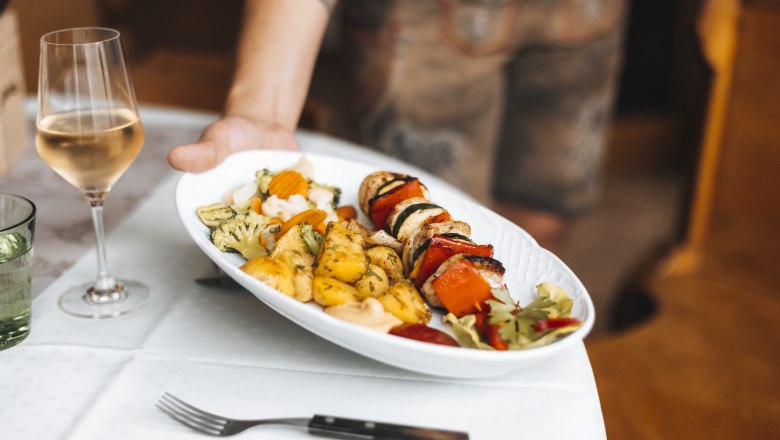 Ein Teller mit Grillspieß, Gemüse und Kartoffeln wird serviert, daneben ein Glas Weißwein.