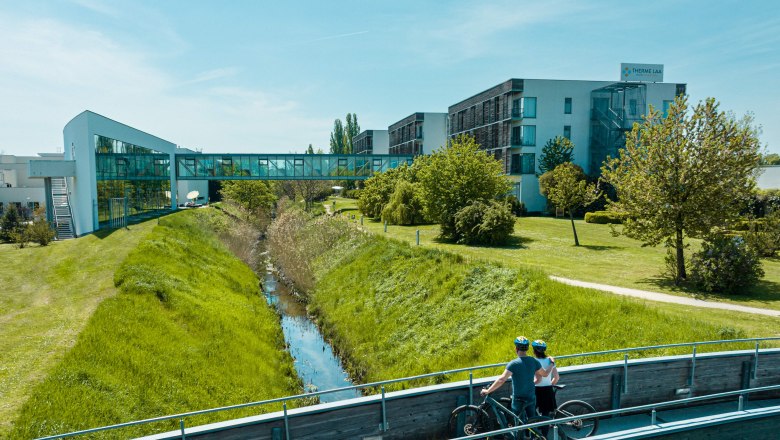 Zwei Radfahrer auf einer Br&uuml;cke vor der Therme Laa & Hotel.