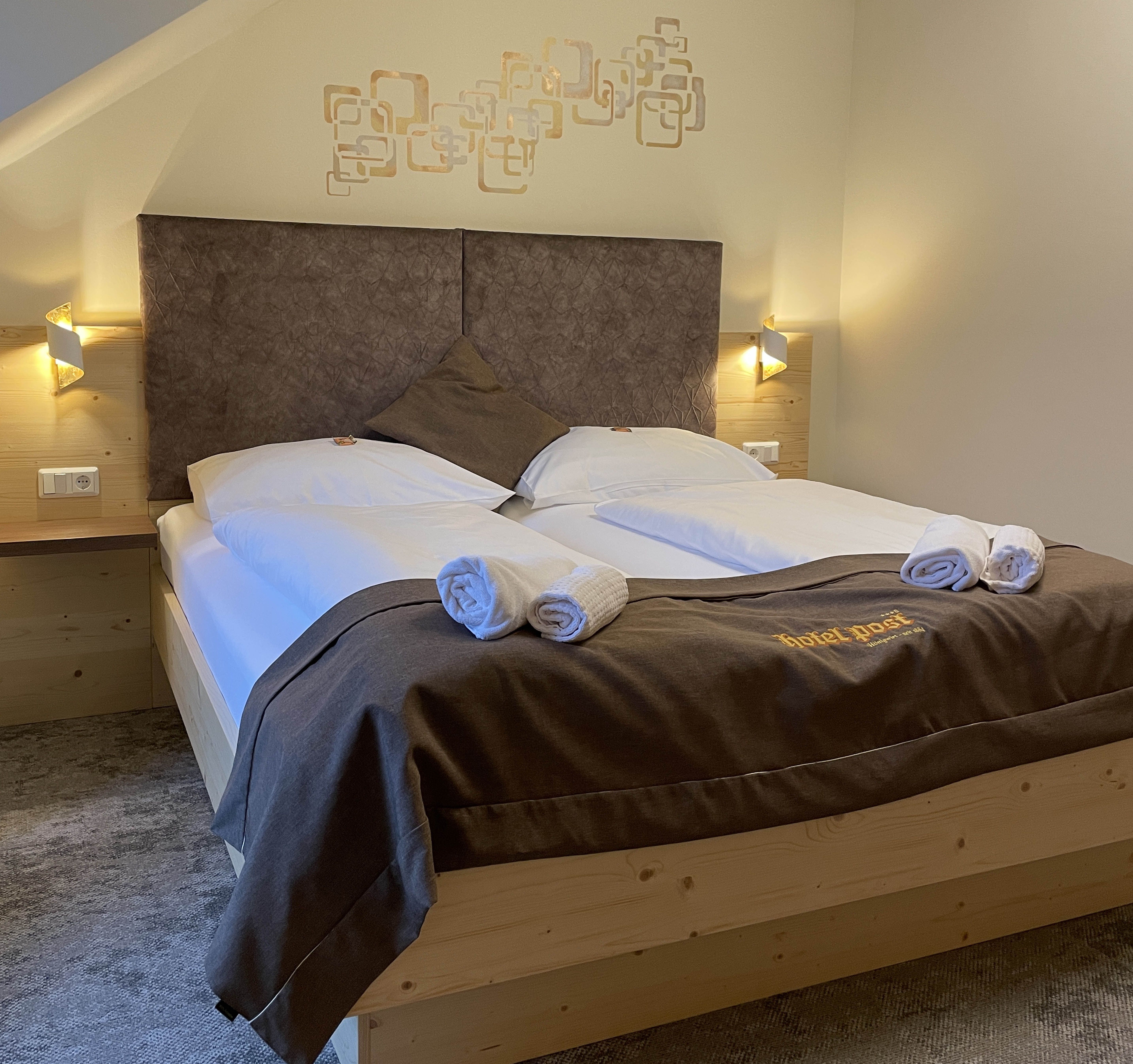 Ein gemütliches Doppelzimmer mit großem Bett, dekoriert mit brauner Bettwäsche und Handtüchern. An der Wand sind geometrische Muster.