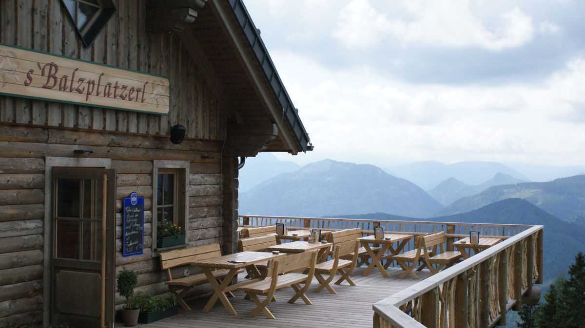 Holzterrasse mit Tischen und Bänken vor einer Berghütte mit Blick auf die Alpen.